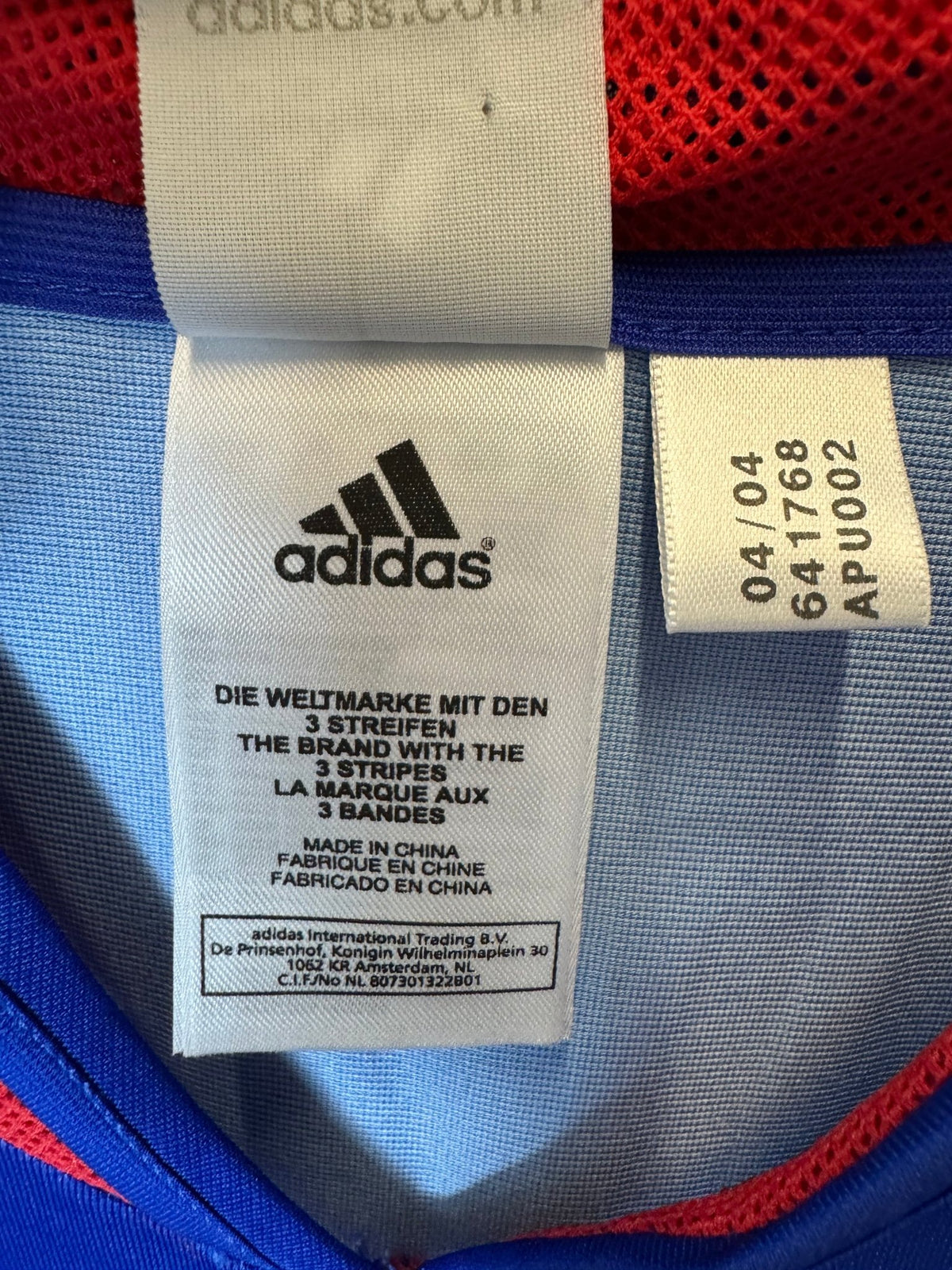 Close-up of an Adidas garment interior label with the Adidas logo and size/lot information, shown against blue and red mesh fabric.