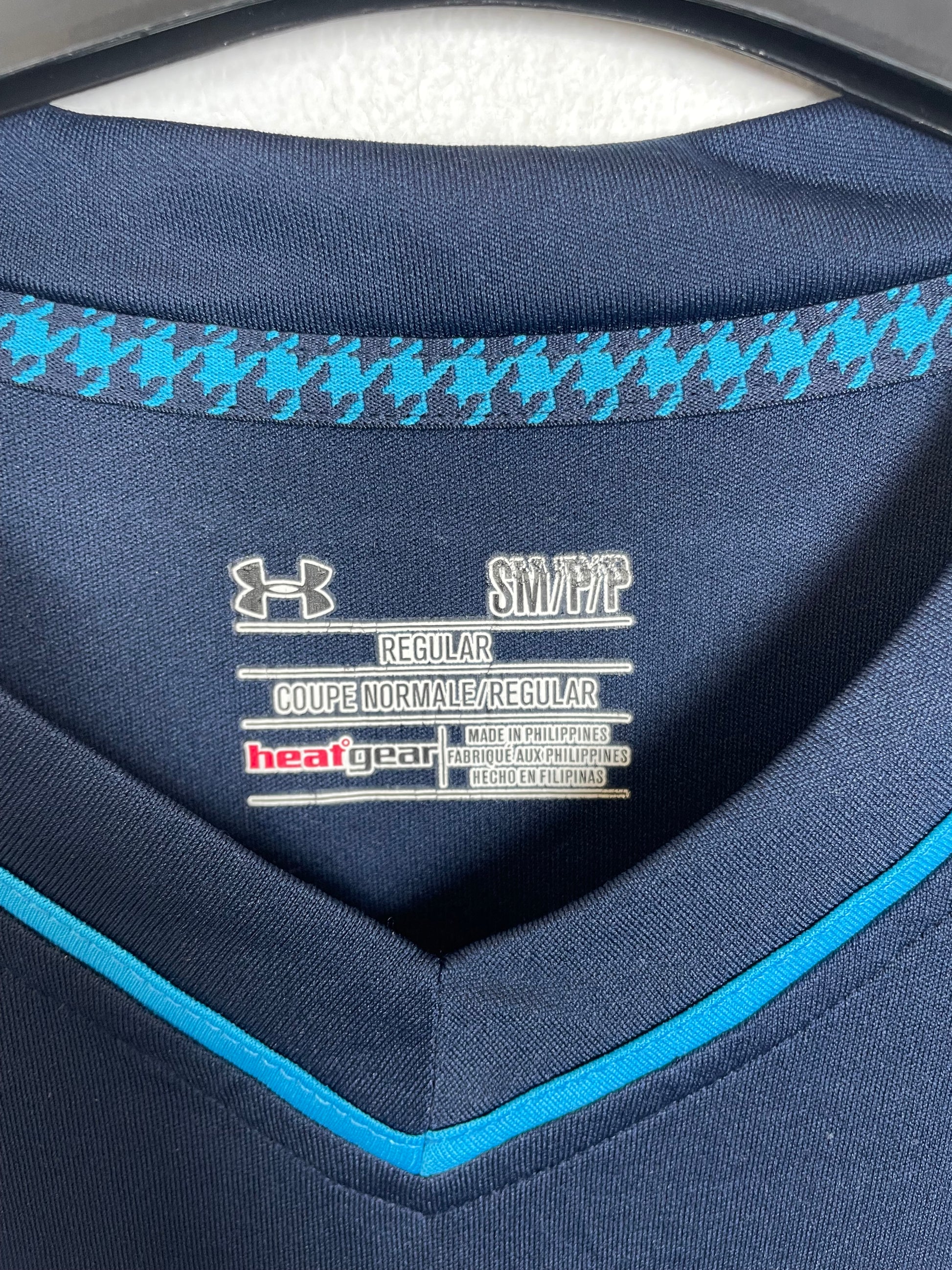 Close-up of the inside collar of a navy blue Under Armour athletic top, showing the inner label with 'SWEAT' and 'REGULAR' text, plus a light blue houndstooth pattern along the inner neckline.