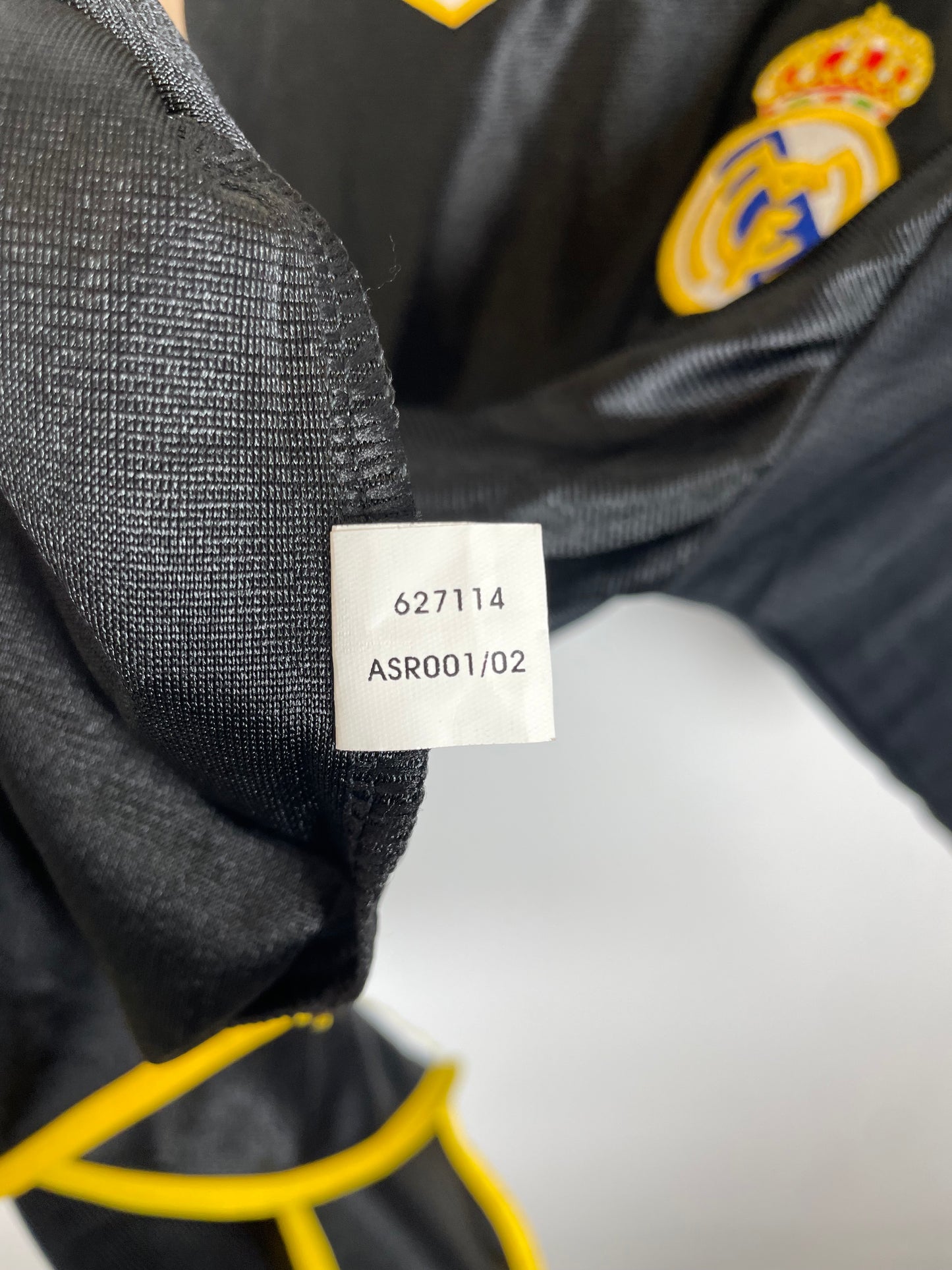 Close-up of a black Real Madrid soccer jersey showing the white sewn-in size tag with printed numbers and the club's embroidered crest on the chest.