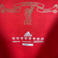 Close-up of a red fabric surface showing a gold-toned Manchester United crest and text, plus a white Adidas logo and care labels on the right.