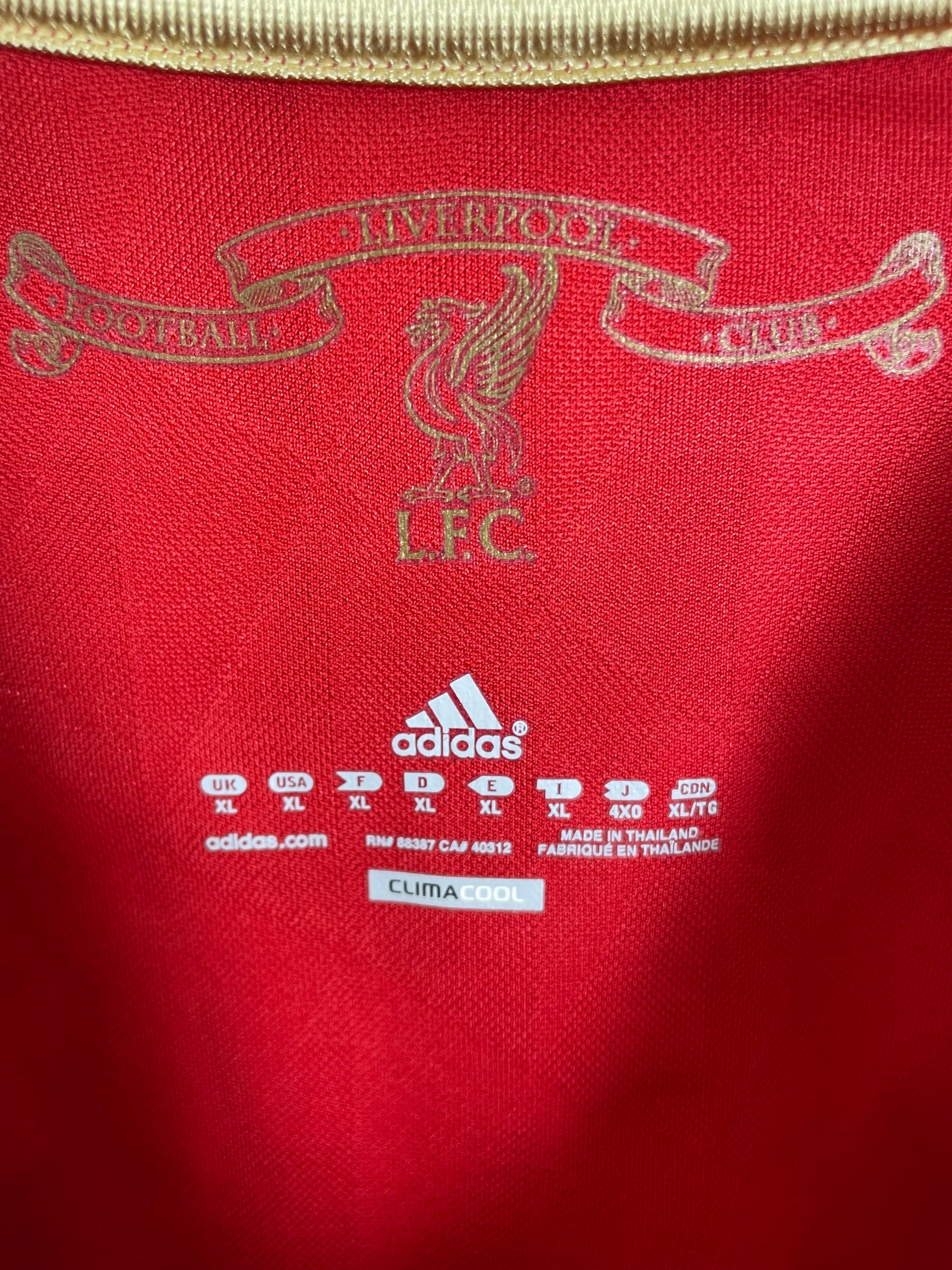 Close-up of a red fabric surface showing a gold-toned Manchester United crest and text, plus a white Adidas logo and care labels on the right.
