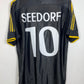 A black Adidas soccer jersey with 'SEEDORF' and the number '10' printed in white on the back, featuring yellow stripes on the sleeves, hanging on a wooden hanger against a white wall.