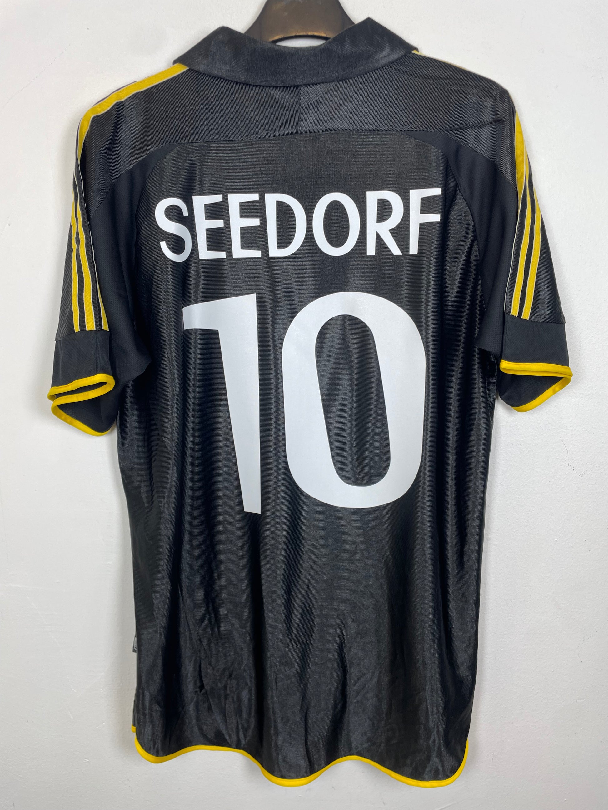 A black Adidas soccer jersey with 'SEEDORF' and the number '10' printed in white on the back, featuring yellow stripes on the sleeves, hanging on a wooden hanger against a white wall.