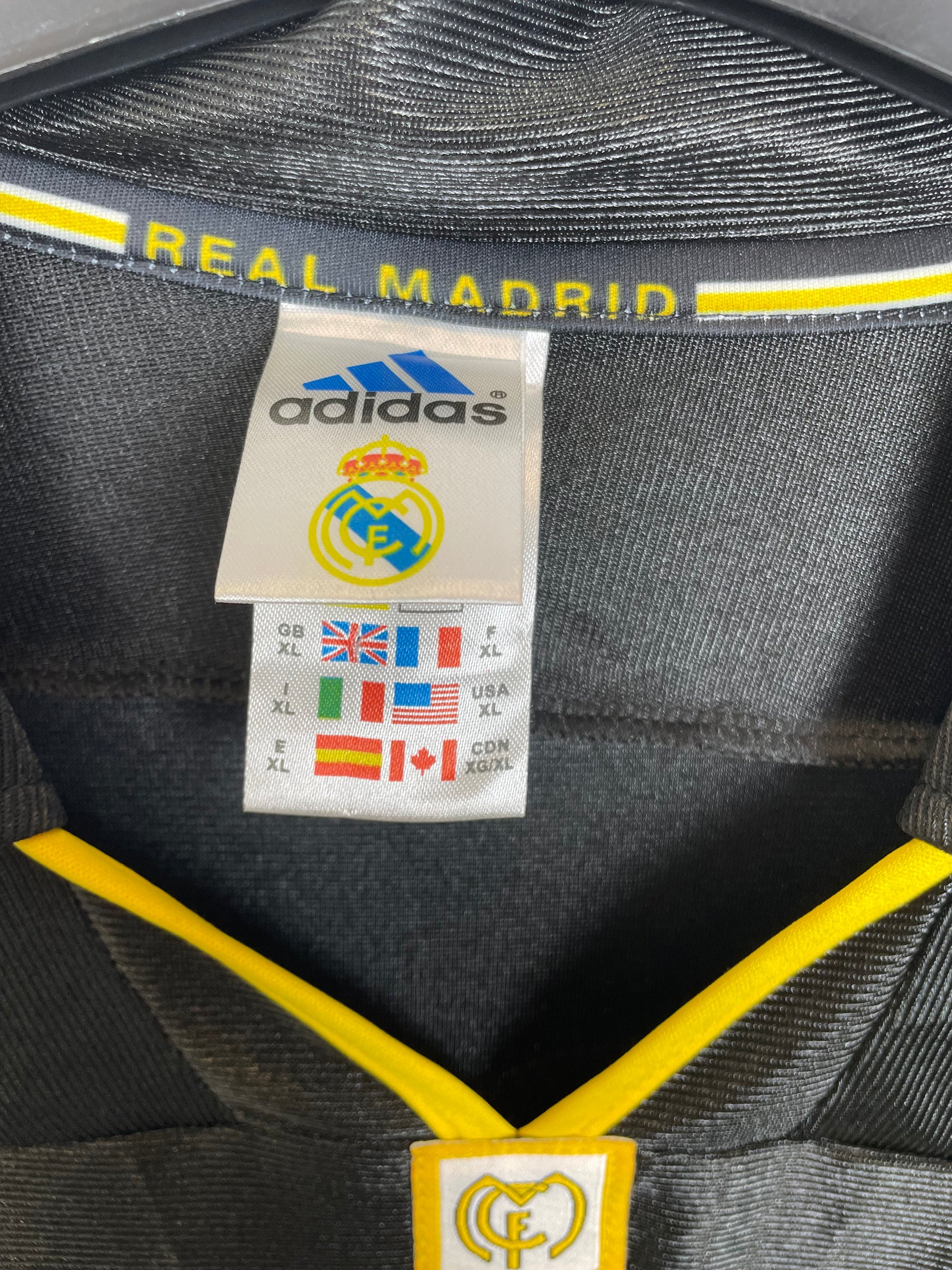 Close-up of the interior label of a black Adidas Real Madrid bag, showing the brand logo, team crest, and size information, with yellow accents and the text 'REAL MADRID' on the side.