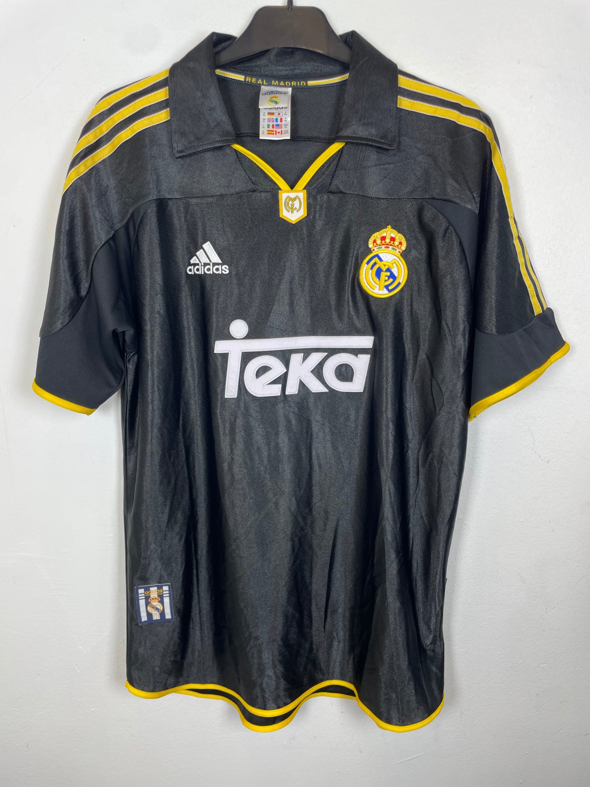 A black Real Madrid soccer jersey with yellow trim and white logos, laid flat on a light gray background.